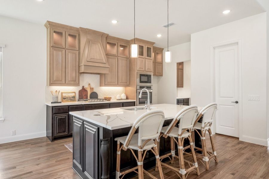Kitchen with dark island, white quartz countertop, light wood cabinets, custom range hood, and three bar stools on hardwood floors