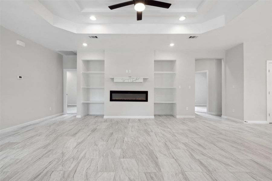 Unfurnished living room with a raised ceiling, built in features, a ceiling fan, a glass covered fireplace, and recessed lighting Unfurnished living room with a raised ceiling, built in features, a ceiling fan, a glass covered fireplace, and recessed lighting