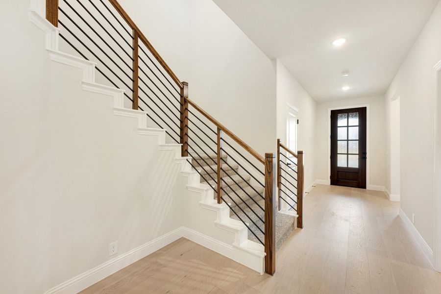 Staircase with wood handrail and black metal balusters in an entryway with light hardwood floors and dark front door Staircase with wood handrail and black metal balusters in an entryway with light hardwood floors and dark front door