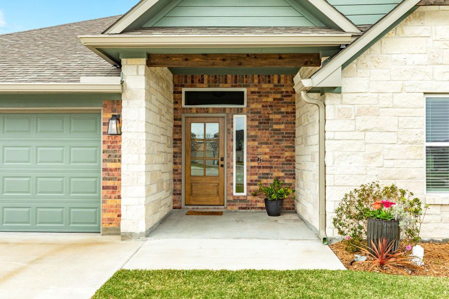 This photo showcases a welcoming front entrance of a modern home. It features a combination of brick and stone facade, a double garage door, and a wooden front door with glass panels. The tidy porch is accented with potted plants, and the well-maintained lawn enhances the curb appeal.