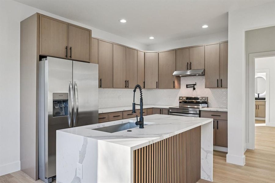 Kitchen with light stone counters, light wood-style floors, stainless steel appliances, decorative backsplash, and an island with sink