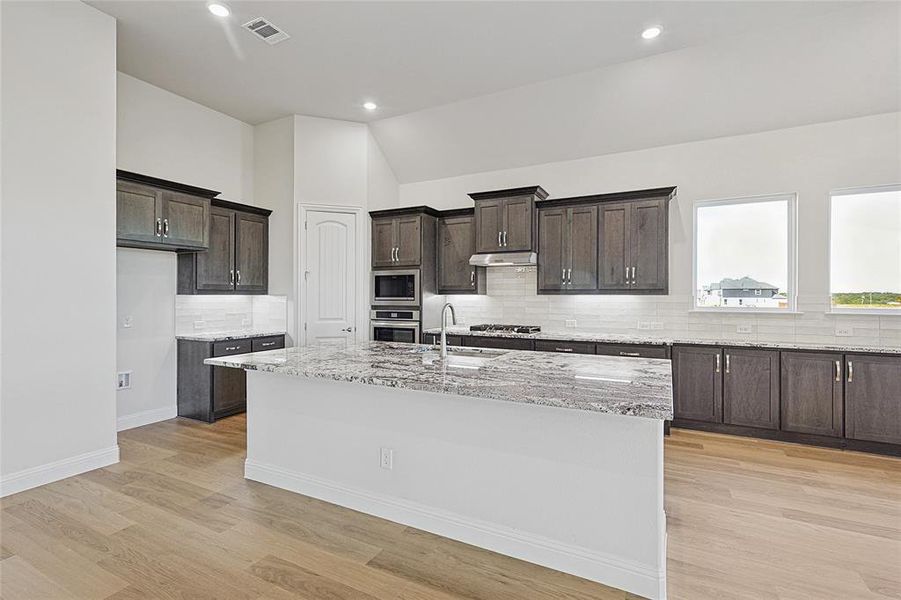Kitchen with light stone countertops, dark wood finish cabinetry, tasteful backsplash, a center island with sink, and light wood-style flooring