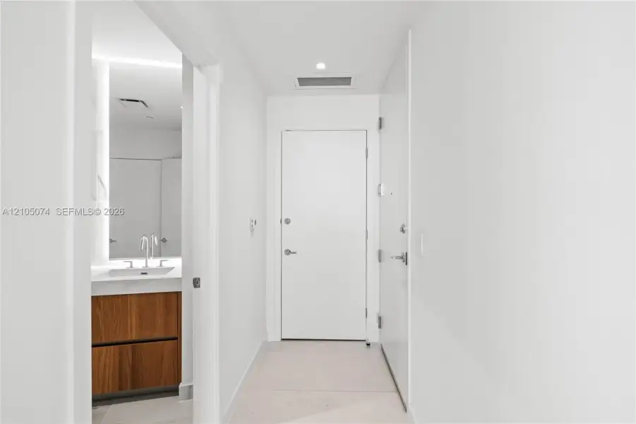 Neutral palette hallway featuring minimalist white interior doors, recessed lighting, and large-format tile flooring, with a glimpse of a wood-finish vanity and integrated basin