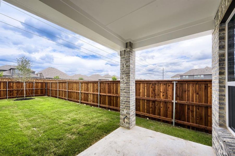 Covered patio featuring brick columns, concrete flooring, and a full privacy fence enclosing a grass lawn with a young tree
