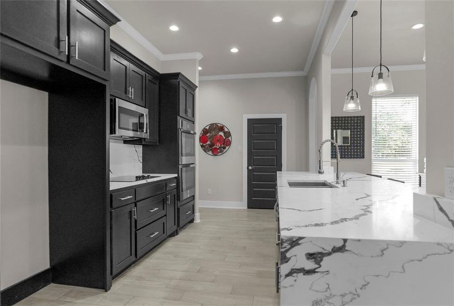 Kitchen featuring light stone counters, ornamental molding, dark cabinets, pendant lighting, and light wood-type flooring