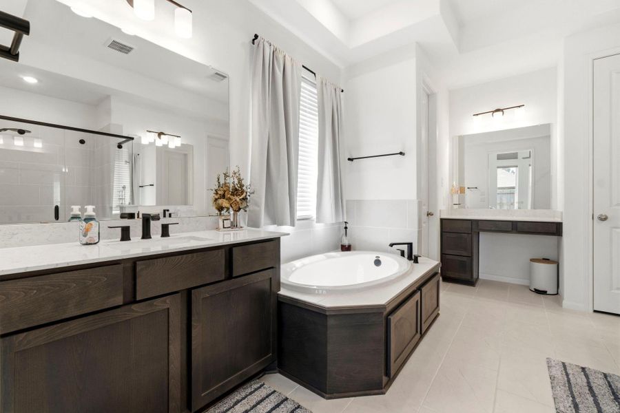 This spacious bathroom features a luxurious soaking tub, dual vanities with dark wood cabinets, and bright natural lighting. A large mirror and a separate makeup area add functionality and elegance.