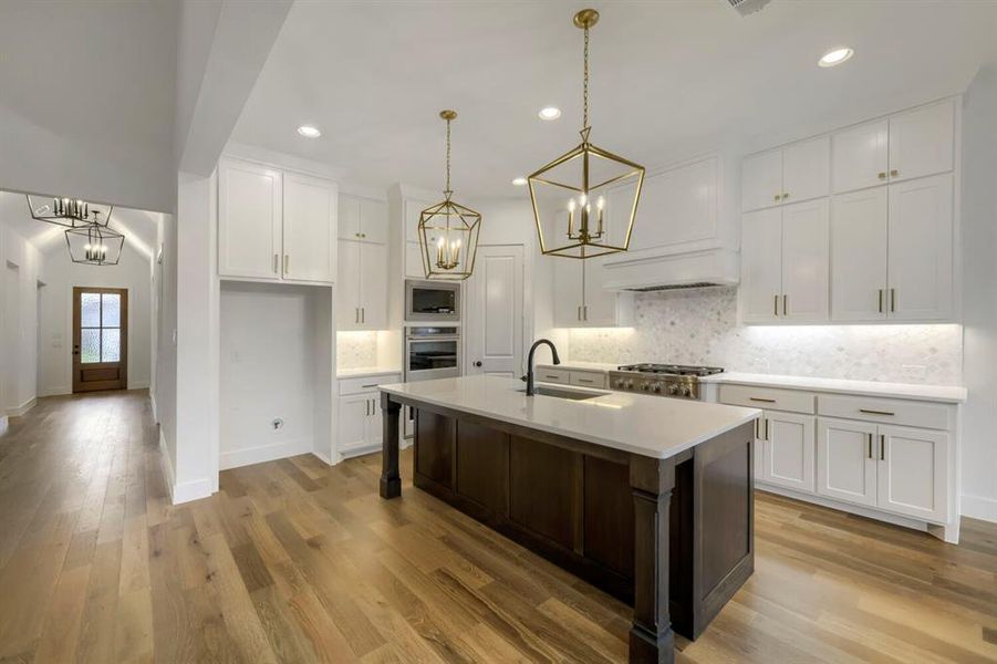 The kitchen features a large island with a countertop, white cabinetry with gold-toned hardware, a tile backsplash, and hardwood floors