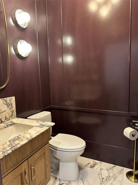 Bathroom featuring panel accent wall, natural stone vanity top, light wood-finish vanity with gold-tone hardware, white commode, and decorative wall sconces