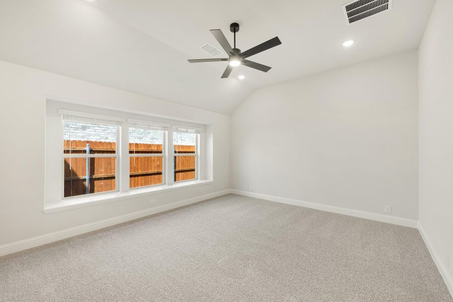 Empty bedroom with vaulted ceiling, ceiling fan, carpet flooring, and triple window overlooking backyard fence