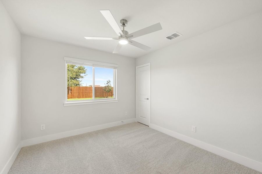 Empty bedroom with white walls, carpet flooring, ceiling fan, and a window overlooking a fenced backyard