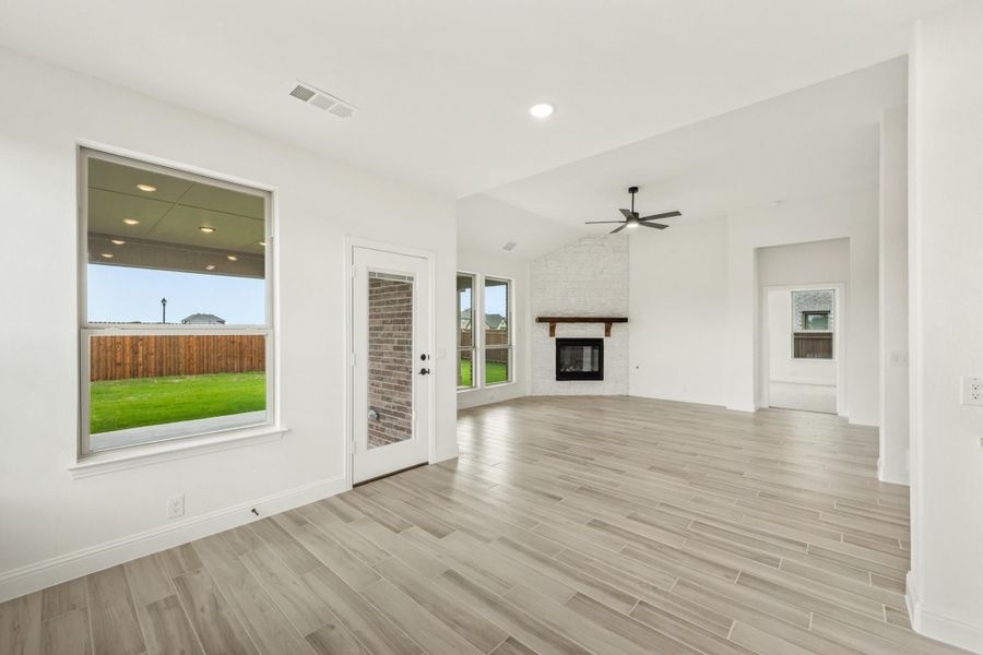 Open living room with light wood floors, brick fireplace, ceiling fan, and backyard view through windows Open living room with light wood floors, brick fireplace, ceiling fan, and backyard view through windows