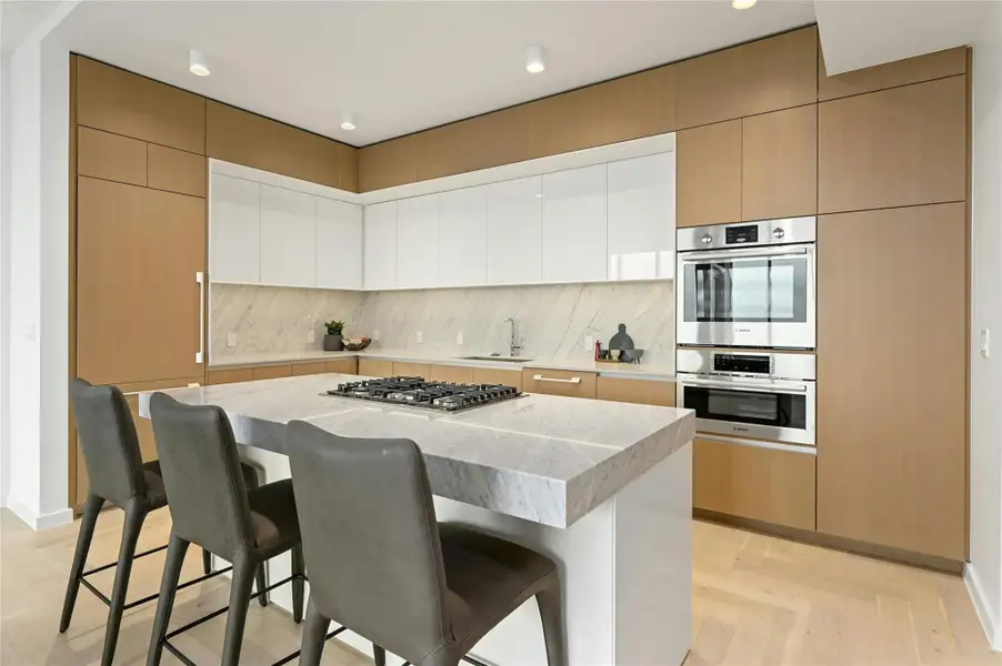 A modern, open-concept kitchen, dining and living area. The kitchen features a sleek, minimalist design with a paneled refrigerator, freezer & dishwasher and reads more like furniture or cabinetry. The island at its center is topped with white-colored Silestone®. The island includes a built-in gas cooktop and stainless steel oven (Photo from previous model Residence No. 2104 on lower level)