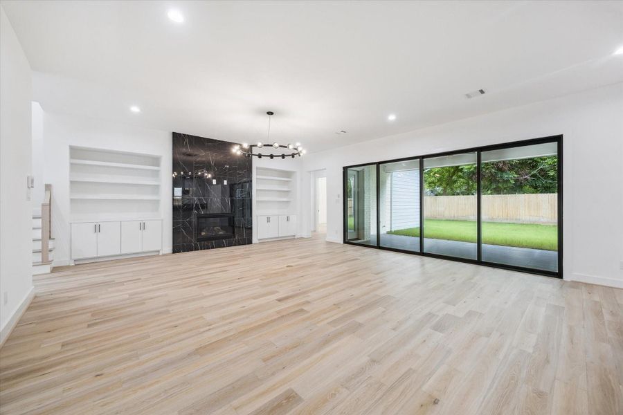 Expansive and bright, the living room features a dramatic black marble fireplace flanked by custom built-ins, wide-plank floors, and a statement light fixture. A full wall of sliding glass doors opens to the covered patio, creating a seamless indoor-outdoor connection perfect for entertaining. Expansive and bright, the living room features a dramatic black marble fireplace flanked by custom built-ins, wide-plank floors, and a statement light fixture. A full wall of sliding glass doors opens to the covered patio, creating a seamless indoor-outdoor connection perfect for entertaining.