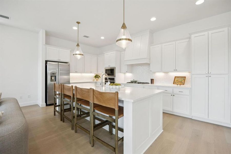 Spacious kitchen featuring an expansive central island with seating, white shaker cabinetry, stainless steel appliances, pendant lighting, and light wood-finish flooring