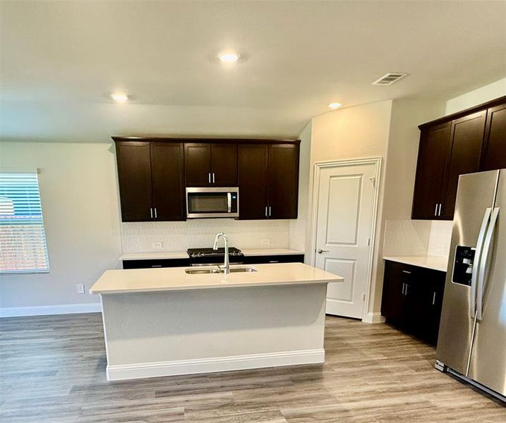 Modern kitchen featuring dark wood cabinetry, light countertops, stainless steel appliances, a center island with sink, and wood-finish flooring Modern kitchen featuring dark wood cabinetry, light countertops, stainless steel appliances, a center island with sink, and wood-finish flooring