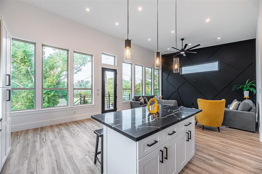 This modern kitchen features sleek white cabinetry with a contrasting black island, high ceilings, and ample natural light from large windows. It opens to a cozy living area with stylish accents, creating a seamless space for entertaining.