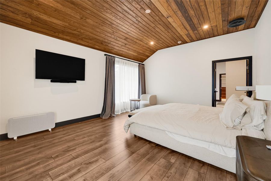 Wood-paneled ceiling and elegant wood flooring. It includes a wall-mounted TV, large windows with curtains for natural light, and a cozy seating area. The room is modern and inviting, perfect for relaxation.