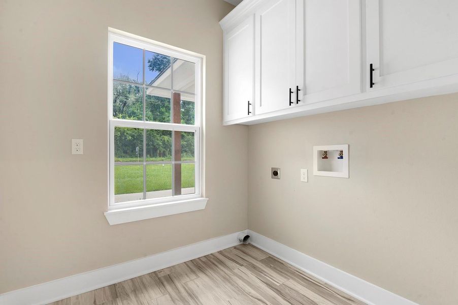This is a bright laundry room featuring large windows, white cabinets with black handles, and hookups for a washer and dryer. The room has light-colored walls and flooring, creating a clean and inviting space with a view of greenery outside. This is a bright laundry room featuring large windows, white cabinets with black handles, and hookups for a washer and dryer. The room has light-colored walls and flooring, creating a clean and inviting space with a view of greenery outside.