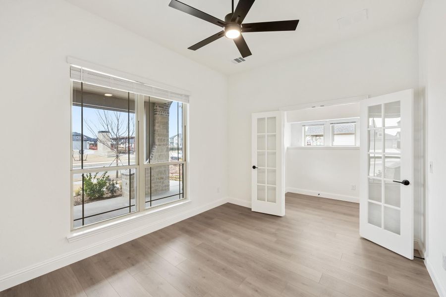 Empty room with hardwood floors, ceiling fan, large window, and French doors leading to adjacent space Empty room with hardwood floors, ceiling fan, large window, and French doors leading to adjacent space