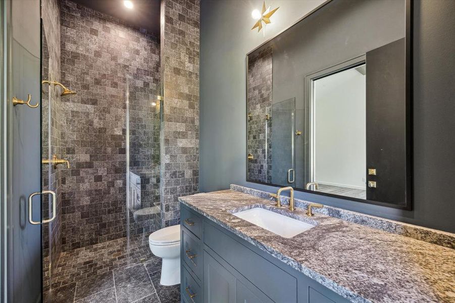 This bathroom exudes a sophisticated, dark aesthetic with a richly textured stone tile shower and seamless shower glass. The sleek vanity is topped with a matching stone countertop, and the brass fixtures throughout add a touch of warmth. Accessible from bedroom as well as from a second door that leads to the exterior outdoor patio/kitchen. With the additional access/layout of the bathroom it creates convenience and functionality to make it ideal to be a future pool bath.