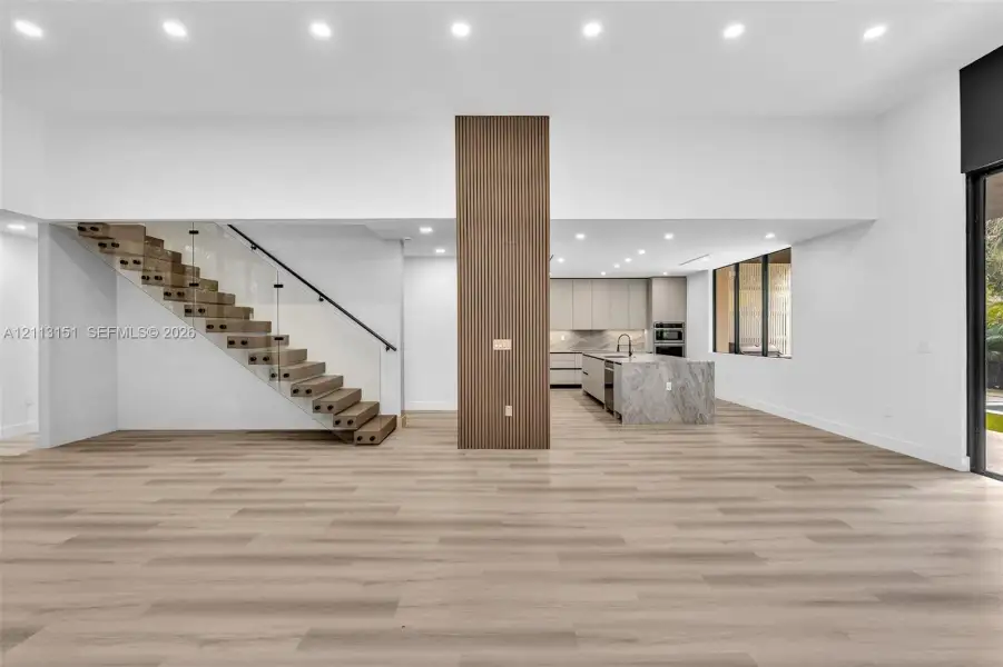 Open-concept interior featuring wood-finish flooring, a central floor-to-ceiling slatted wood accent pillar, recessed lighting, and a staircase with glass railings and wood treads Open-concept interior featuring wood-finish flooring, a central floor-to-ceiling slatted wood accent pillar, recessed lighting, and a staircase with glass railings and wood treads