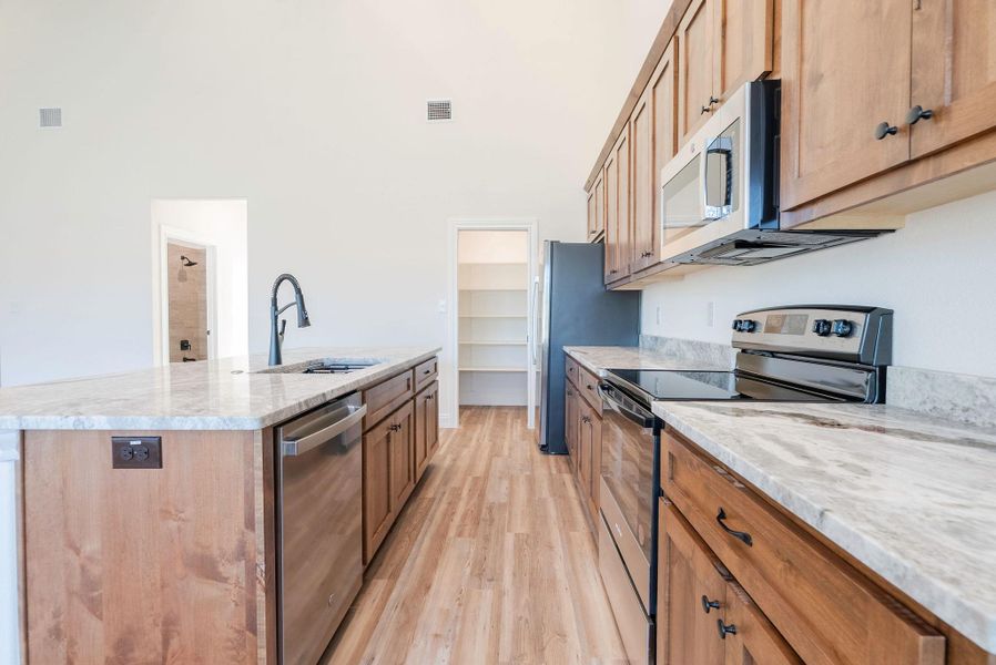 Kitchen featuring stainless steel appliances, light wood-style floors, light stone counters, a kitchen island with sink, and brown cabinetry Kitchen featuring stainless steel appliances, light wood-style floors, light stone counters, a kitchen island with sink, and brown cabinetry