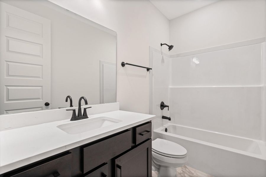 Modern bathroom featuring a dark wood-finish vanity with a white countertop and integrated sink, matte black hardware, an expansive wall mirror, and a white tub/shower combination with a matching matte black shower head and faucet Modern bathroom featuring a dark wood-finish vanity with a white countertop and integrated sink, matte black hardware, an expansive wall mirror, and a white tub/shower combination with a matching matte black shower head and faucet