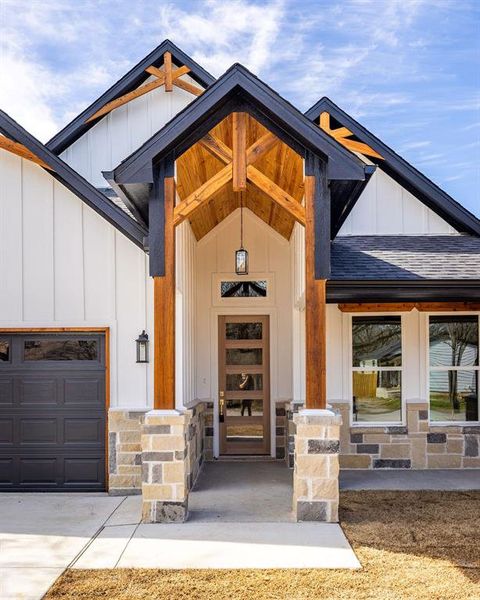 View of exterior entry featuring concrete driveway, stone siding, roof with shingles, an attached garage, and board and batten siding