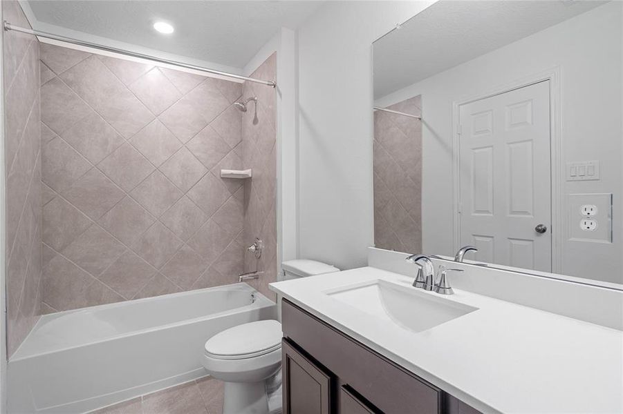 Secondary bath features tile flooring, bath/shower combo with tile surround, stained wood cabinets, beautiful light countertops, mirror, sleek fixtures and modern finishes.