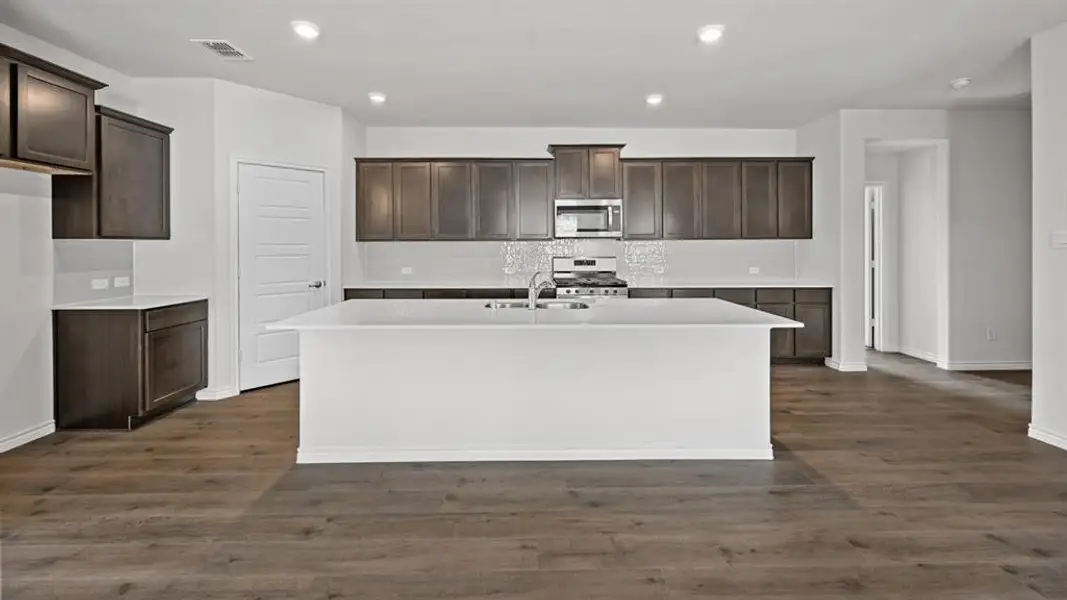 Open-concept kitchen featuring a white center island with integrated sink, dark wood-finish cabinetry, white countertops, a mosaic tile backsplash, and stainless steel appliances Open-concept kitchen featuring a white center island with integrated sink, dark wood-finish cabinetry, white countertops, a mosaic tile backsplash, and stainless steel appliances