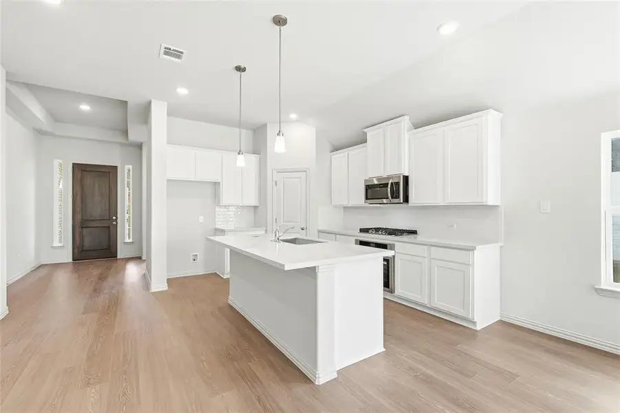 Kitchen with white cabinetry, light wood finished floors, hanging light fixtures, an island with sink, and stainless steel appliances Kitchen with white cabinetry, light wood finished floors, hanging light fixtures, an island with sink, and stainless steel appliances