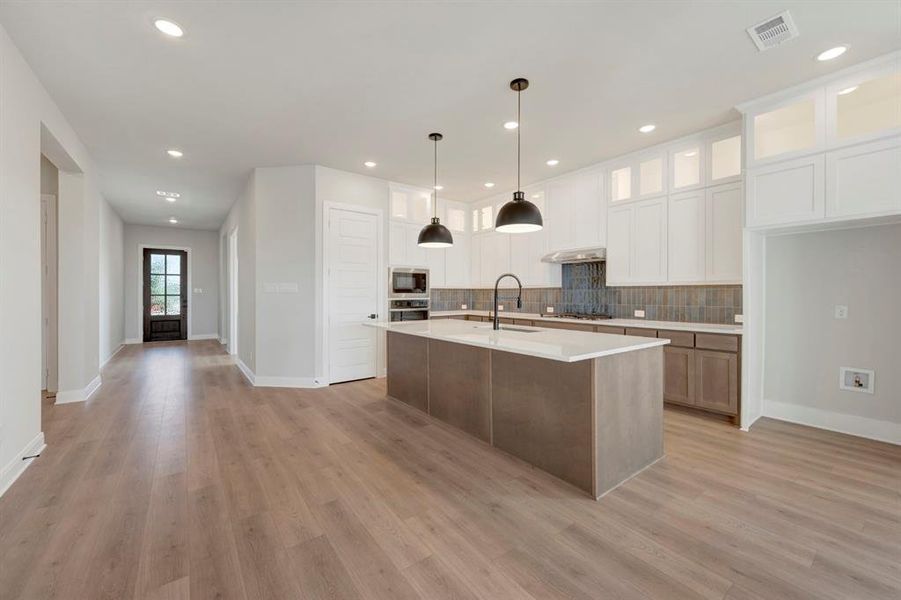 Kitchen with two tone cabinetry, a center island with sink, decorative light fixtures, glass insert cabinets, and tasteful backsplash
