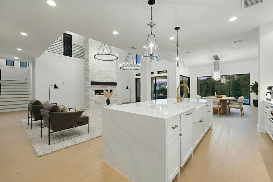 Open-concept living space featuring a central kitchen island with a white countertop, light wood-finish flooring, a stacked stone fireplace, and large windows