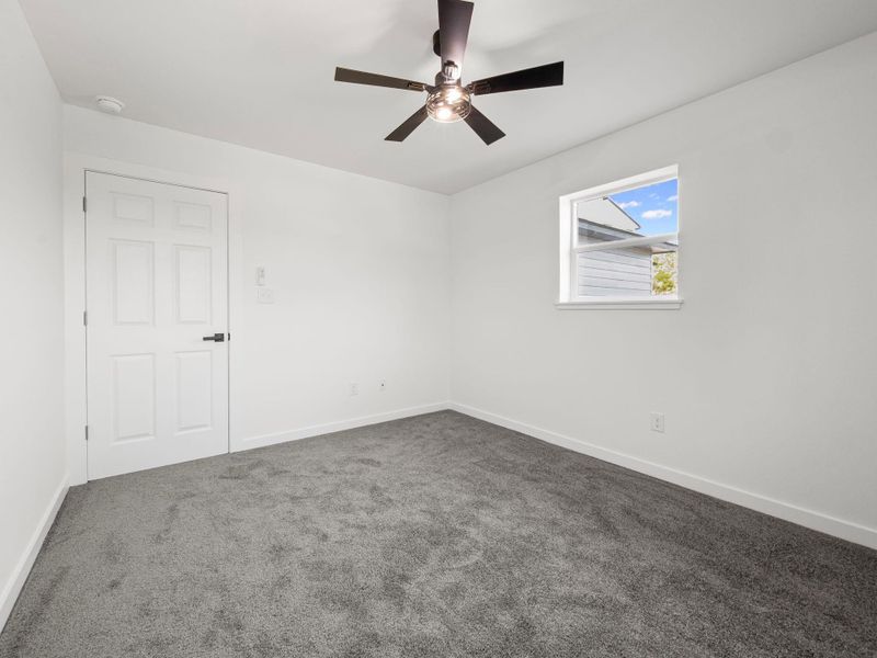 Bright room featuring soft gray carpeting, a multi-paneled white door with dark hardware, a modern ceiling fan, white baseboards, and a window providing natural light