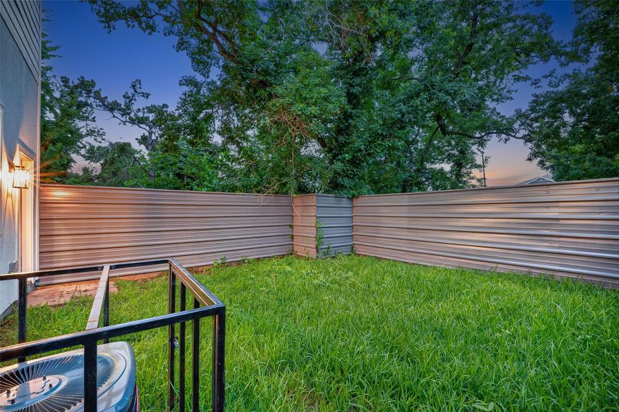 This photo shows a small, fenced backyard with lush grass and surrounded by trees, offering privacy and a tranquil atmosphere. A central air conditioning unit is visible, indicating modern amenities. This photo shows a small, fenced backyard with lush grass and surrounded by trees, offering privacy and a tranquil atmosphere. A central air conditioning unit is visible, indicating modern amenities.