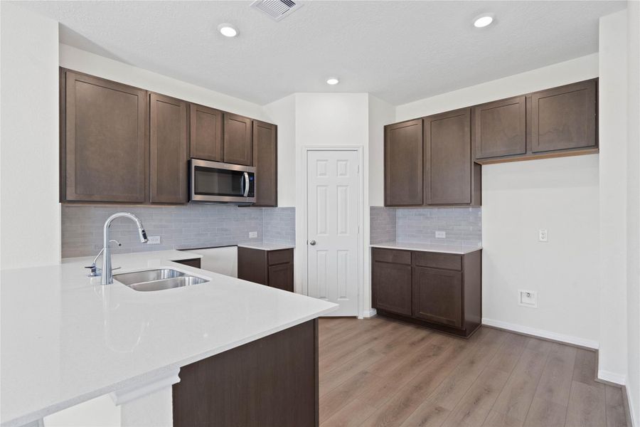 The kitchen boasts high ceilings, recessed lighting, Woodmont Dakota cabinets in a Smolder stain, Silestone countertops in Blanco Maple, stainless steel Whirlpool appliances, a modern Daltile backsplash, an extended breakfast bar countertop, and a pantry—all overlooking the expansive Great Room.