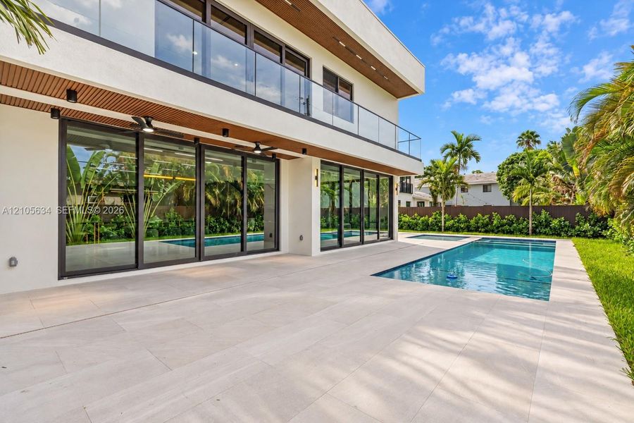 Modern exterior featuring a wrap-around glass balcony, oversized floor-to-ceiling sliding glass doors, a stone-tiled patio, and an in-ground swimming pool