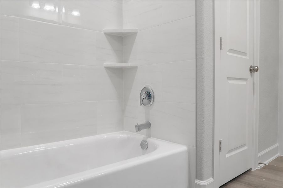 This photo showcases a modern bathroom featuring a clean, white bathtub with sleek fixtures and a tiled wall. There's a corner shelf for storage and a closed door with a polished knob on the side, adding to the room's contemporary style.