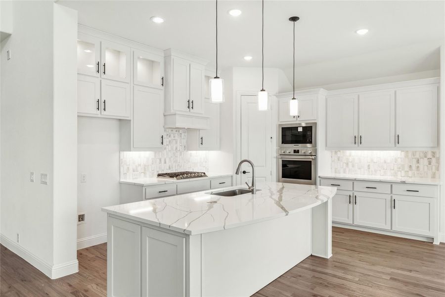 White kitchen with large marble island, white cabinets, pendant lights, and stainless steel double ovens on hardwood floors.