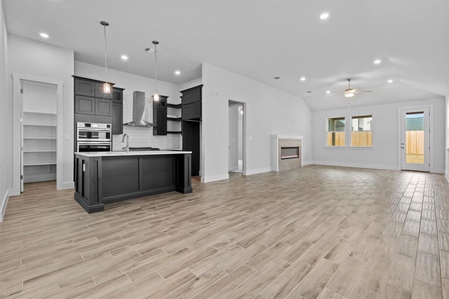 Kitchen featuring ceiling fan, decorative backsplash, a kitchen island with sink, wall chimney range hood, and pendant lighting