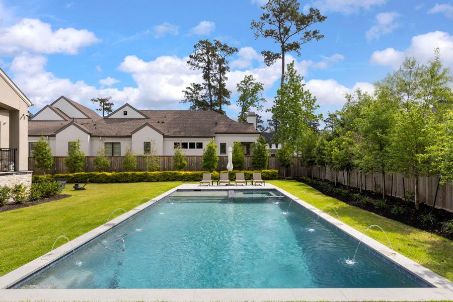 Resort-style backyard featuring a stunning pool with water features, expansive green space, and mature trees creating a private setting perfect for relaxing or entertaining outdoors.