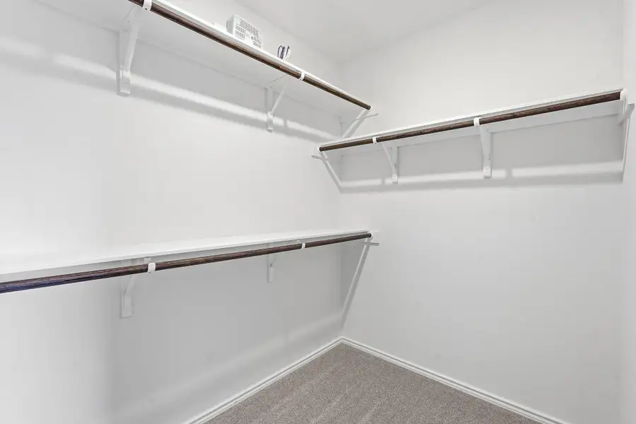 Organized closet space featuring white shelving, dark-finish hanging rods, and light gray carpet flooring Organized closet space featuring white shelving, dark-finish hanging rods, and light gray carpet flooring