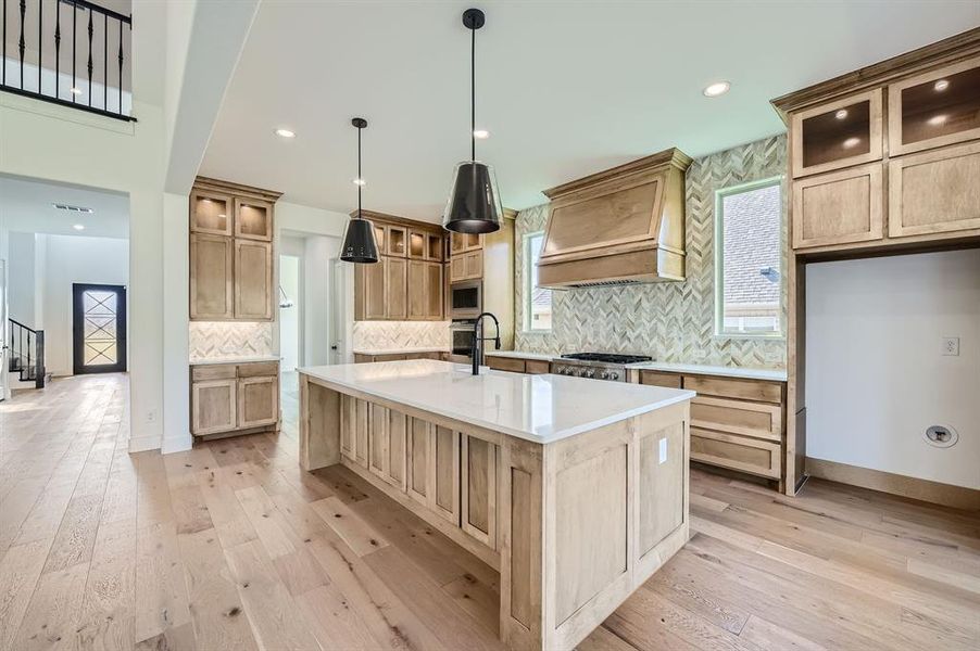 Kitchen with tasteful backsplash, an island with sink, light hardwood / wood-style floors, decorative light fixtures, and custom exhaust hood