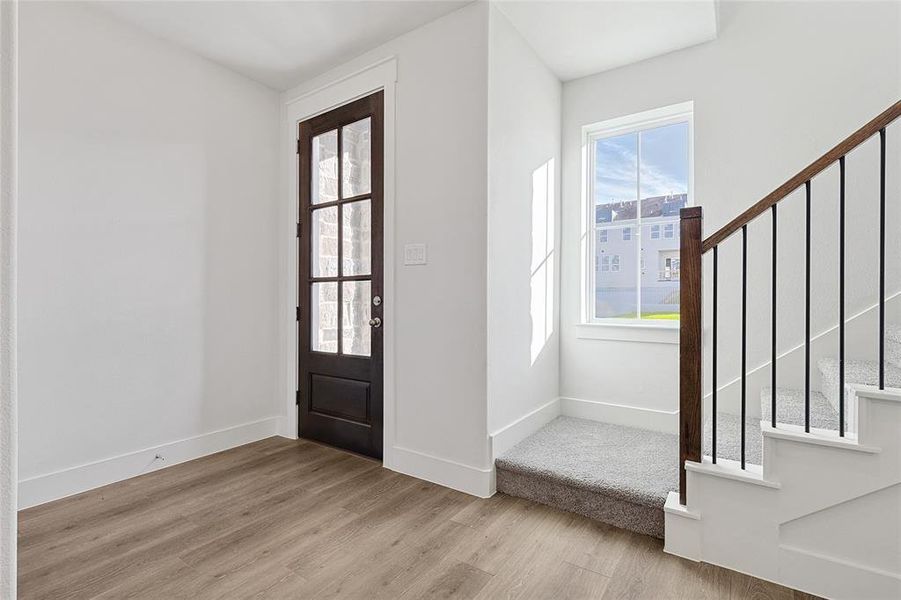 The entryway features wood-look flooring and a dark wood door with glass paneling, complemented by a staircase with carpeted steps and a dark wood handrail with black balusters