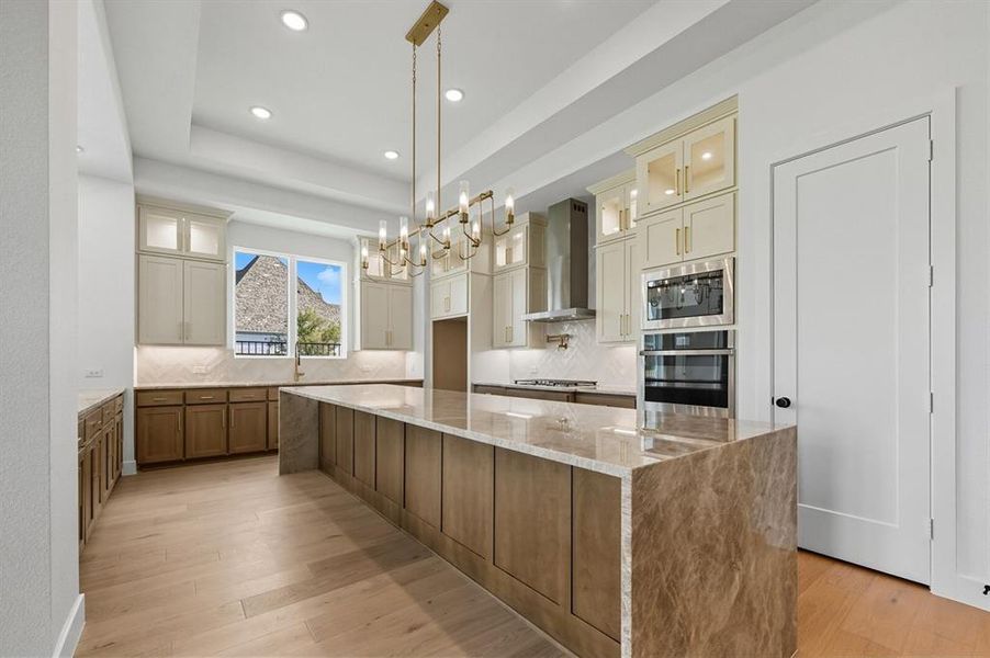 Kitchen with glass insert cabinets, tasteful backsplash, light stone counters, a large island, and brown cabinets Kitchen with glass insert cabinets, tasteful backsplash, light stone counters, a large island, and brown cabinets