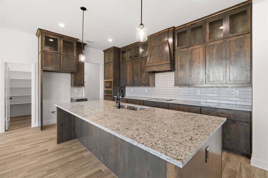 Kitchen with decorative backsplash, light stone countertops, dark brown cabinetry, a kitchen island with sink, and hanging light fixtures Kitchen with decorative backsplash, light stone countertops, dark brown cabinetry, a kitchen island with sink, and hanging light fixtures