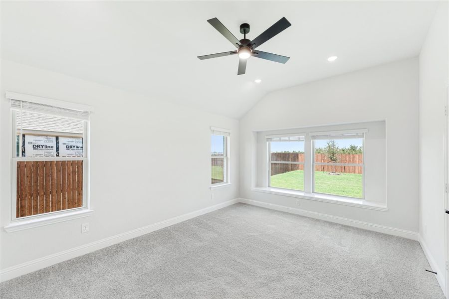 Empty bedroom with vaulted ceiling, ceiling fan, carpet flooring, and multiple windows overlooking backyard