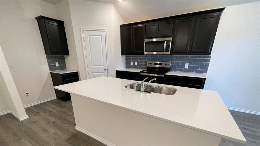 Kitchen featuring a central island with an undermount double-basin stainless steel sink, white countertops, dark cabinetry, stainless steel appliances, and grey subway tile backsplash