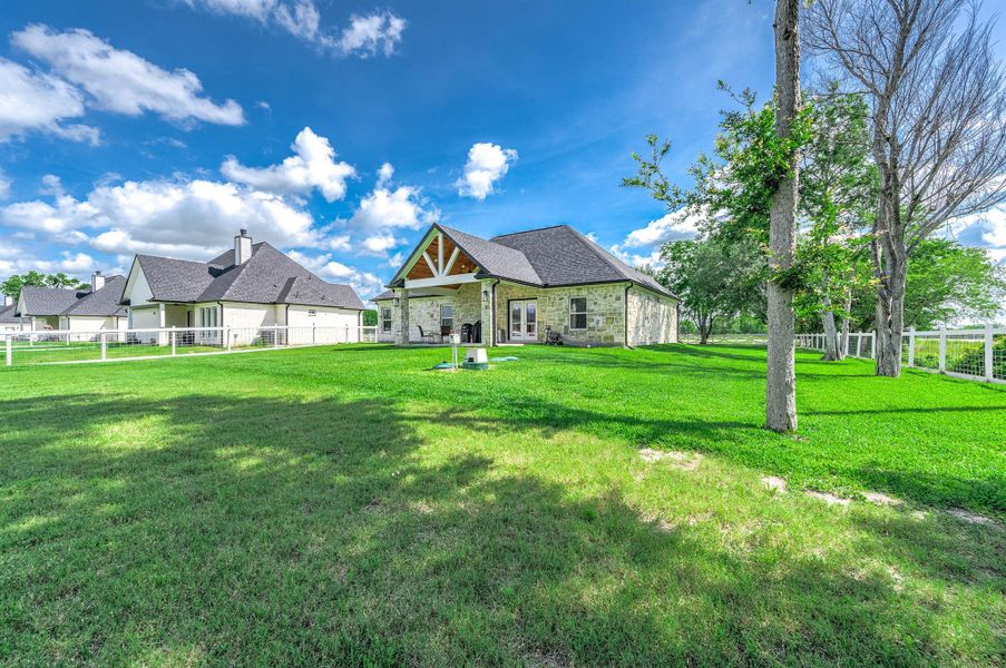 This view highlights the home’s beautiful stone exterior, spacious yard, and peaceful setting surrounded by open green space and mature trees. The covered patio and fenced backyard add to the property’s charm and outdoor appeal.