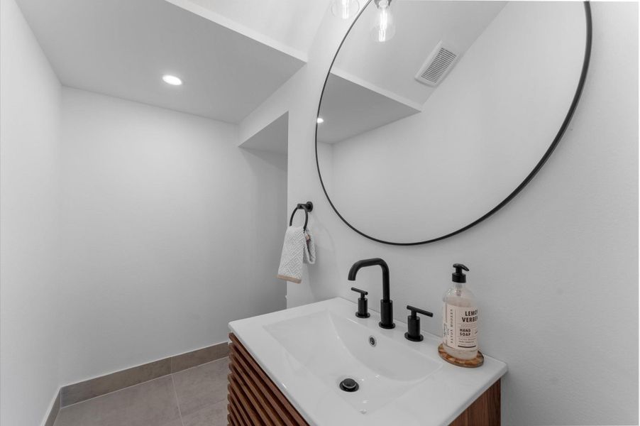 This stylish first-floor powder room blends modern elegance with functional design. Featuring contemporary finishes throughout, the space includes a sleek vanity with clean lines, minimalist hardware, and a polished countertop. The vanity is paired with a statement mirror and bold lighting fixtures that add visual interest.